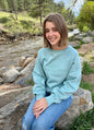 Person wearing a light blue sweatshirt sitting by a river with trees in the background